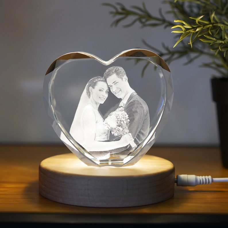 May include: A heart-shaped crystal with a 3D engraved image of a couple, illuminated by a warm light on a wooden base.