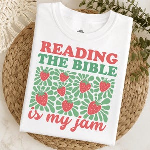 May include: White t-shirt with the text "READING THE BIBLE is my jam" in red and green. The design includes strawberries and green floral accents, creating a playful and faith-based theme.