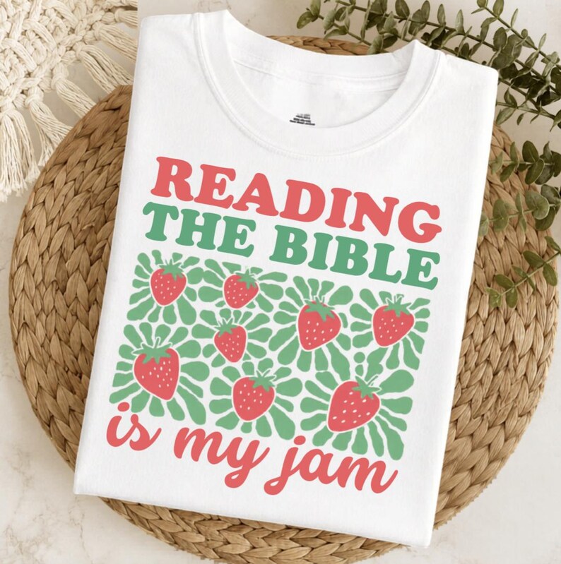 May include: White t-shirt with the text "READING THE BIBLE is my jam" in red and green. The design includes strawberries and green floral accents, creating a playful and faith-based theme.