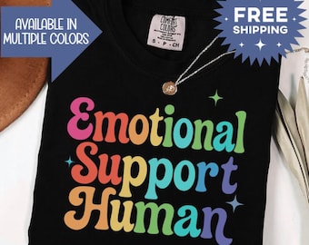 May include: Black t-shirt with the words "Emotional Support Human" in rainbow colors. The shirt has a crew neck and short sleeves. A silver necklace with a small pendant is visible. The shirt is labeled "Comfort Colors" and is available in multiple colors.