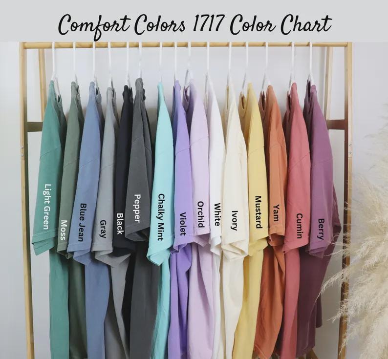 Op de afbeelding: Een kleurenkaart van Comfort Colors 1717 t-shirts, weergegeven op een houten rek. De shirts zijn in verschillende kleuren, waaronder lichtgroen, spijkerbroekblauw, grijs, zwart, violet en mosterd. De tekst "Comfort Colors 1717 Color Chart" staat bovenaan.