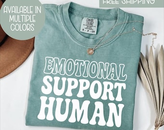 May include: A teal Comfort Colors t-shirt with the words "EMOTIONAL SUPPORT HUMAN" in white, retro-style lettering. The shirt has a crew neck and is accessorized with a silver necklace. The text "AVAILABLE IN MULTIPLE COLORS" is in a circle in the upper left.