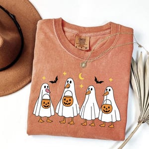 May include: A terracotta-colored Comfort Colors t-shirt featuring a Halloween-themed design. The graphic includes four ghost-like figures holding jack-o'-lantern buckets, with bats, stars, and a crescent moon above them. A gold necklace and a brown hat are also visible.