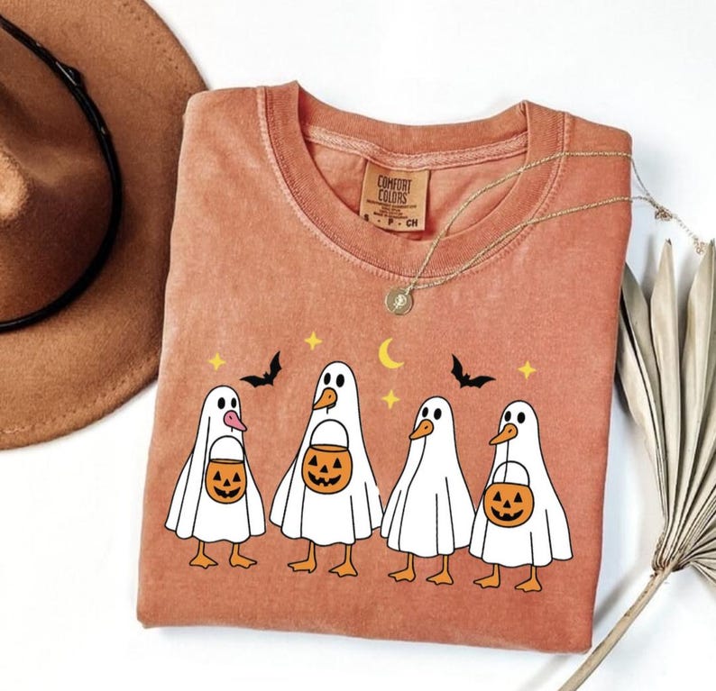 May include: A terracotta-colored Comfort Colors t-shirt featuring a Halloween-themed design. The graphic includes four ghost-like figures holding jack-o'-lantern buckets, with bats, stars, and a crescent moon above them. A gold necklace and a brown hat are also visible.