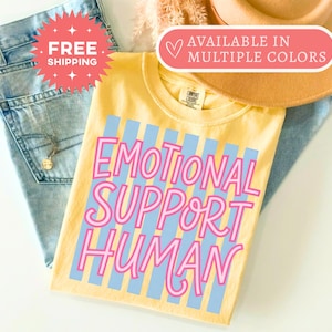 May include: A pale yellow t-shirt featuring a graphic design of vertical light blue stripes and the text "EMOTIONAL SUPPORT HUMAN" written in a bold, pink, rounded font. The shirt has a crew neckline and a visible brand tag at the inner collar.