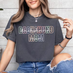 May include: Dark gray t-shirt with the text "DACHSHUND MOM" in a colorful, outlined font. The shirt is a crew neck style and appears to be made of a soft material. The text is in a rainbow gradient.