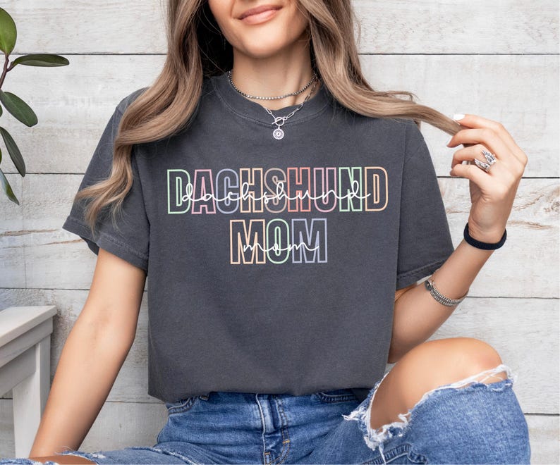 May include: Dark gray t-shirt with the text "DACHSHUND MOM" in a colorful, outlined font. The shirt is a crew neck style and appears to be made of a soft material. The text is in a rainbow gradient.