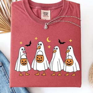 May include: A maroon t-shirt featuring a Halloween-themed graphic. The design includes four cartoon ducks dressed as ghosts, each holding a jack-o'-lantern bucket. The shirt also has bats, stars, and a crescent moon. A silver necklace is draped across the shirt.