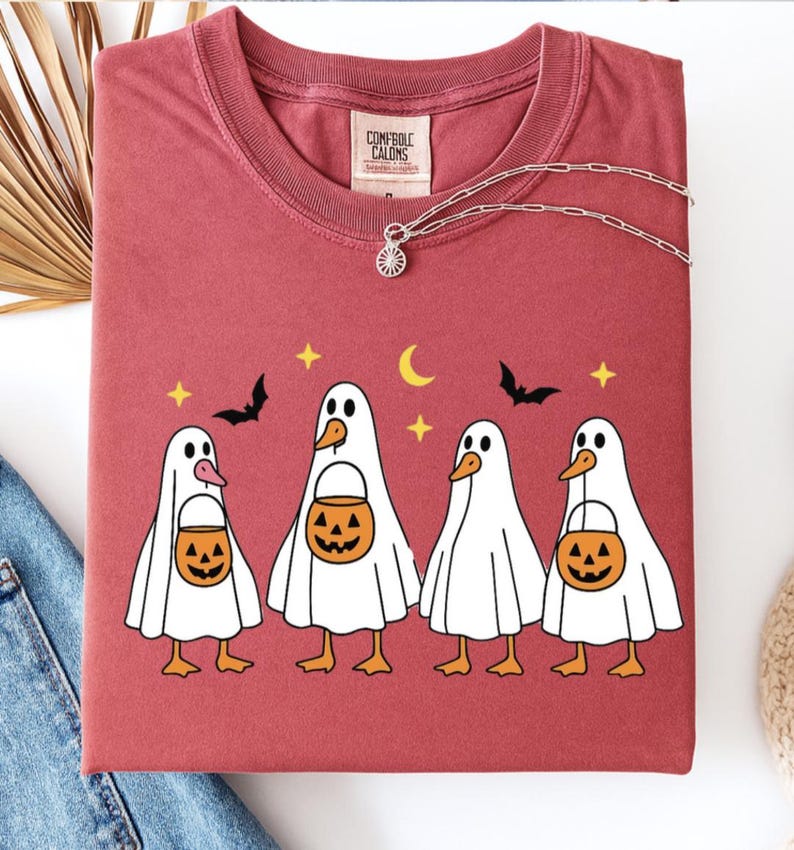 May include: A maroon t-shirt featuring a Halloween-themed graphic. The design includes four cartoon ducks dressed as ghosts, each holding a jack-o'-lantern bucket. The shirt also has bats, stars, and a crescent moon. A silver necklace is draped across the shirt.