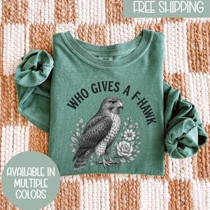 May include: A green long-sleeve shirt with the text "WHO GIVES A F-HAWK" surrounding a detailed illustration of a hawk. The shirt also has a floral design. The shirt is available in multiple colors.
