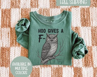 May include: A green long-sleeve shirt with the text "HOO GIVES A F-" and a detailed illustration of an owl perched on a branch. The shirt is a solid color and has rolled-up sleeves. The phrase "AVAILABLE IN MULTIPLE COLORS" is in a circle at the bottom.