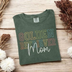 K&ouml;nnte beinhalten: Ein gr&uuml;nes T-Shirt mit dem Schriftzug "GOLDEN RETRIEVER Mom" in bunten Blockbuchstaben und Schrift. Das Shirt liegt gefaltet auf einer Holzoberfl&auml;che, mit getrockneten Pflanzen und Blumen im Hintergrund.