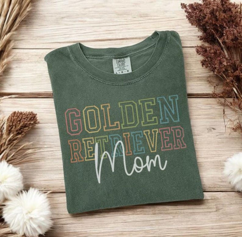 K&ouml;nnte beinhalten: Ein gr&uuml;nes T-Shirt mit dem Schriftzug "GOLDEN RETRIEVER Mom" in bunten Blockbuchstaben und Schrift. Das Shirt liegt gefaltet auf einer Holzoberfl&auml;che, mit getrockneten Pflanzen und Blumen im Hintergrund.