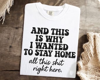 May include: White t-shirt with the text "AND THIS IS WHY I WANTED TO STAY HOME all this shit right here" in black, displayed on a wooden surface. The shirt is folded and has a crew neck.