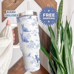 May include: A white tumbler with a handle and a straw, featuring a blue and white cat toile pattern. The words "STRAW INCLUDED" are visible. The tumbler is held by a person.