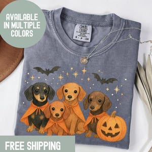 Op de afbeelding: Een grijs Comfort Colors T-shirt met een Halloween-ontwerp. Het heeft vier teckels met oranje capes, vleermuizen, sterren en een pompoen. Het shirt heeft de tekst "AVAILABLE IN MULTIPLE COLORS" en een zilveren ketting.