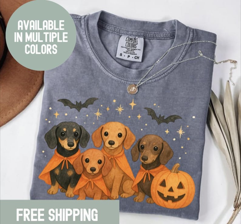 Op de afbeelding: Een grijs Comfort Colors T-shirt met een Halloween-ontwerp. Het heeft vier teckels met oranje capes, vleermuizen, sterren en een pompoen. Het shirt heeft de tekst "AVAILABLE IN MULTIPLE COLORS" en een zilveren ketting.