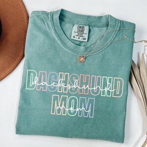 May include: A teal Comfort Colors t-shirt with the words "DACHSHUND MOM" in a colorful, outlined font. A silver necklace with a small pendant rests on the shirt. The shirt is folded on a white surface.