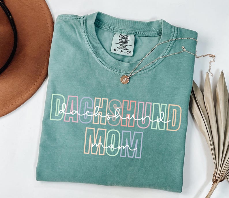 May include: A teal Comfort Colors t-shirt with the words "DACHSHUND MOM" in a colorful, outlined font. A silver necklace with a small pendant rests on the shirt. The shirt is folded on a white surface.