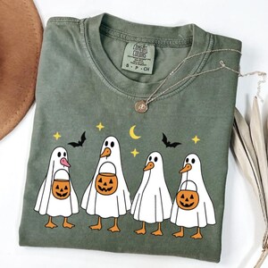 May include: Olive green Comfort Colors t-shirt featuring a Halloween-themed graphic. The design includes five cartoon ghost-like figures with duck-like features, each holding a jack-o'-lantern bucket. Bats, stars, and a crescent moon complete the festive design.