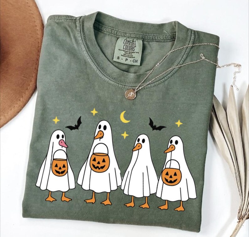 May include: Olive green Comfort Colors t-shirt featuring a Halloween-themed graphic. The design includes five cartoon ghost-like figures with duck-like features, each holding a jack-o'-lantern bucket. Bats, stars, and a crescent moon complete the festive design.