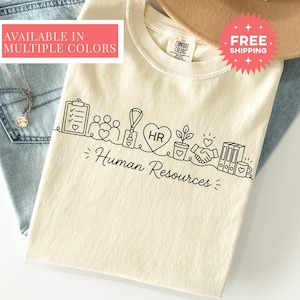 May include: A cream-colored short-sleeve t-shirt featuring a black line art graphic of various office icons including a clipboard, a group of people, an ID badge, a heart with the letters HR, a potted plant, a handshake, and binders. The text Human Resources is printed below the icons in a cursive font.