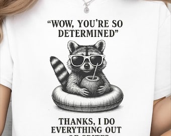 May include: White t-shirt featuring a raccoon wearing sunglasses and an inner tube, holding a drink. The text reads "WOW, YOU'RE SO DETERMINED" and "THANKS, I DO EVERYTHING OUT OF SPITE."