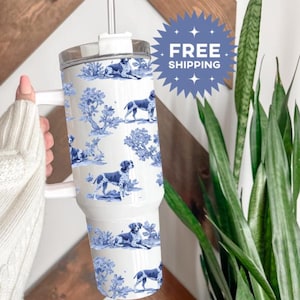 May include: A white tumbler with a blue and white dog and tree pattern. The tumbler has a white handle, a clear lid, and a white straw. The words "FREE SHIPPING" are on a blue starburst.