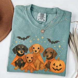 Op de afbeelding: Een turquoise Comfort Colors T-shirt met een Halloween-ontwerp. Het ontwerp bevat vier teckel honden met oranje capes, vleermuizen, sterren en een uitgeholde pompoen. Een zilveren ketting met een hanger is ook zichtbaar.