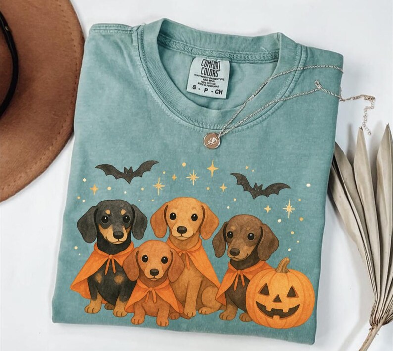 Op de afbeelding: Een turquoise Comfort Colors T-shirt met een Halloween-ontwerp. Het ontwerp bevat vier teckel honden met oranje capes, vleermuizen, sterren en een uitgeholde pompoen. Een zilveren ketting met een hanger is ook zichtbaar.