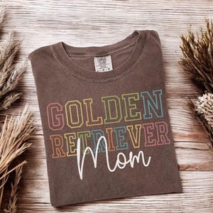 K&ouml;nnte beinhalten: Ein braunes T-Shirt mit dem Aufdruck "GOLDEN RETRIEVER Mom" in bunten Blockbuchstaben. Das Shirt hat einen Rundhalsausschnitt und ein Etikett. Es liegt auf einer Holzoberfl&auml;che mit getrockneten Pflanzen.