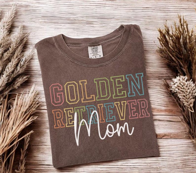K&ouml;nnte beinhalten: Ein braunes T-Shirt mit dem Aufdruck "GOLDEN RETRIEVER Mom" in bunten Blockbuchstaben. Das Shirt hat einen Rundhalsausschnitt und ein Etikett. Es liegt auf einer Holzoberfl&auml;che mit getrockneten Pflanzen.