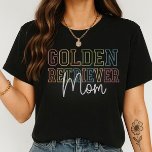 K&ouml;nnte beinhalten: Schwarzes T-Shirt mit dem Schriftzug "GOLDEN RETRIEVER Mom" in einem farbenfrohen, gestapelten Design. Der Text ist in einer Mischung aus Schriftarten und Farben gehalten, wobei das Wort "Mom" handschriftlich gestaltet ist. Das Shirt ist ein l&auml;ssiger Stil.