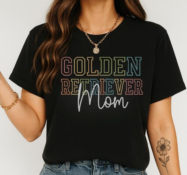 K&ouml;nnte beinhalten: Schwarzes T-Shirt mit dem Schriftzug "GOLDEN RETRIEVER Mom" in einem farbenfrohen, gestapelten Design. Der Text ist in einer Mischung aus Schriftarten und Farben gehalten, wobei das Wort "Mom" handschriftlich gestaltet ist. Das Shirt ist ein l&auml;ssiger Stil.