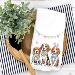 May include: Three beagle dogs wearing Easter-themed costumes and holding Easter eggs. The dogs are wearing bow ties and bunny ears. The background is a white kitchen towel with a colorful banner.