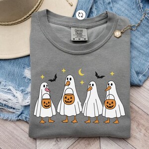 May include: A gray Comfort Colors sweatshirt featuring a Halloween-themed design. The graphic depicts four cartoon ducks dressed as ghosts, each holding a jack-o'-lantern bucket. Bats, stars, and a crescent moon complete the festive design.