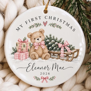 May include: A round white ceramic ornament featuring a brown teddy bear with a pink gingham bow, two wrapped gifts, and a wooden sled. The text reads MY FIRST CHRISTMAS, Eleanor Mae, and 2026, accented with green pine branches and pink bows.
