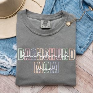May include: A gray sweatshirt with the words "DACHSHUND MOM" in a colorful, outlined font. The sweatshirt is folded on top of a pair of blue denim shorts. A beige hat and gold necklace are also visible.