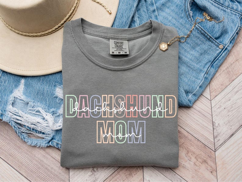 May include: A gray sweatshirt with the words "DACHSHUND MOM" in a colorful, outlined font. The sweatshirt is folded on top of a pair of blue denim shorts. A beige hat and gold necklace are also visible.