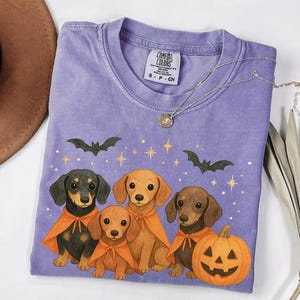 Op de afbeelding: Een paars Comfort Colors T-shirt met een Halloween-thema. De afbeelding toont teckels met oranje capes, vleermuizen, sterren en een uitgesneden pompoen. Een zilveren ketting met een hanger is over het shirt gedrapeerd.