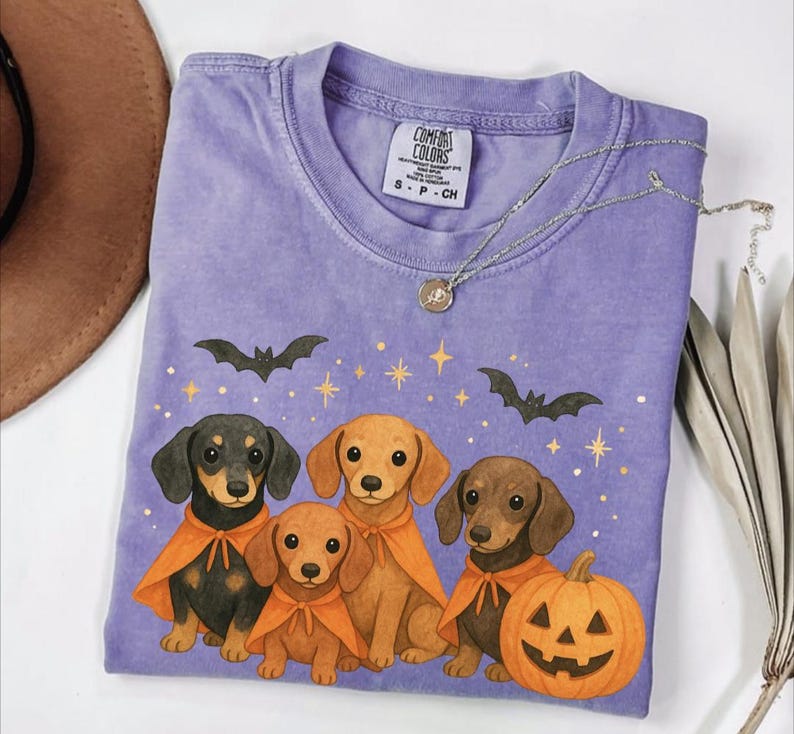 Op de afbeelding: Een paars Comfort Colors T-shirt met een Halloween-thema. De afbeelding toont teckels met oranje capes, vleermuizen, sterren en een uitgesneden pompoen. Een zilveren ketting met een hanger is over het shirt gedrapeerd.