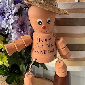 May include: A terracotta pot doll wearing a straw hat, with painted eyes and a red nose. The doll has jointed arms and legs made of small pots. The text "HAPPY GOLDEN ANNIVERSARY" is printed on the doll's body.