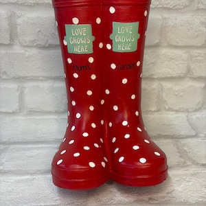 May include: A pair of red wellies with white polka dots. The boots are decorated with green labels that say "Love Grows Here". One boot says "Mum's" and the other says "Garden".