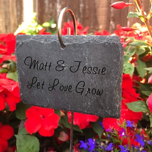 May include: A dark grey slate garden marker with the engraved words "Matt & Jessie Let Love Grow." The marker has a silver metal ring at the top and is set amongst vibrant red and purple flowers.