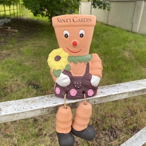 May include: A ceramic garden gnome figurine with a smiling face, holding a yellow sunflower. The gnome is wearing a brown dress with pink polka dots and has black shoes. The gnome is sitting on a metal fence with the words "NANA'S GARDEN" written on its pot.