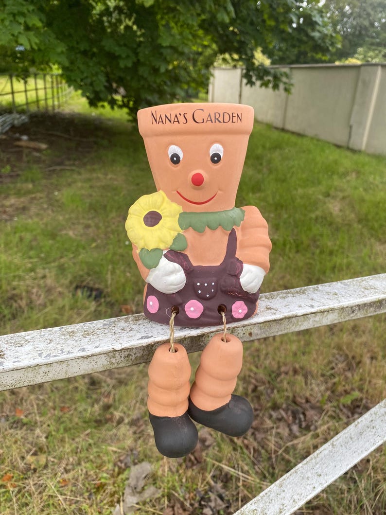 May include: A ceramic garden gnome figurine with a smiling face, holding a yellow sunflower. The gnome is wearing a brown dress with pink polka dots and has black shoes. The gnome is sitting on a metal fence with the words "NANA'S GARDEN" written on its pot.