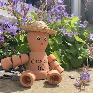 May include: A terracotta garden ornament shaped like a person, with a straw hat and arms and legs made of small pots. The ornament has the word "GRANDAD" and the number "60" printed on the front. It is sitting in front of purple flowers.