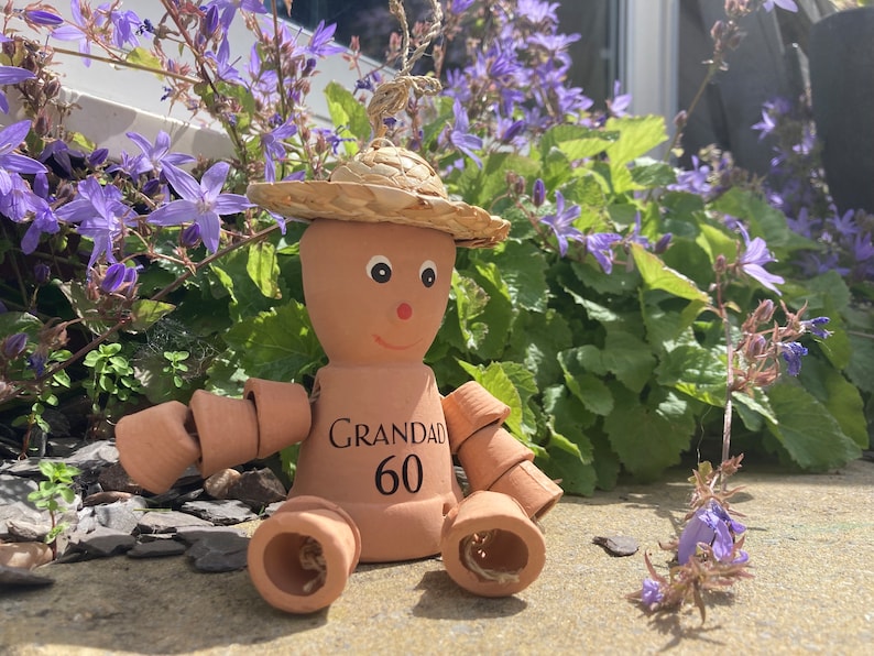 May include: A terracotta garden ornament shaped like a person, with a straw hat and arms and legs made of small pots. The ornament has the word "GRANDAD" and the number "60" printed on the front. It is sitting in front of purple flowers.