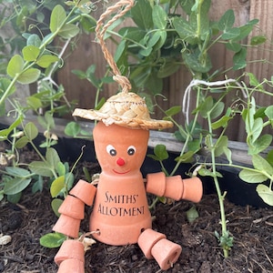 May include: A terracotta garden ornament shaped like a person, wearing a straw hat. The arms and legs are made of small terracotta pots. The text "Smiths Allotment" is visible. The ornament is set amongst green plants.