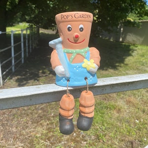 May include: A terracotta garden gnome figurine wearing a blue overall, holding a yellow flower and a blue watering can. The gnome is sitting on a metal fence with the text "Pop's Garden" on the front.